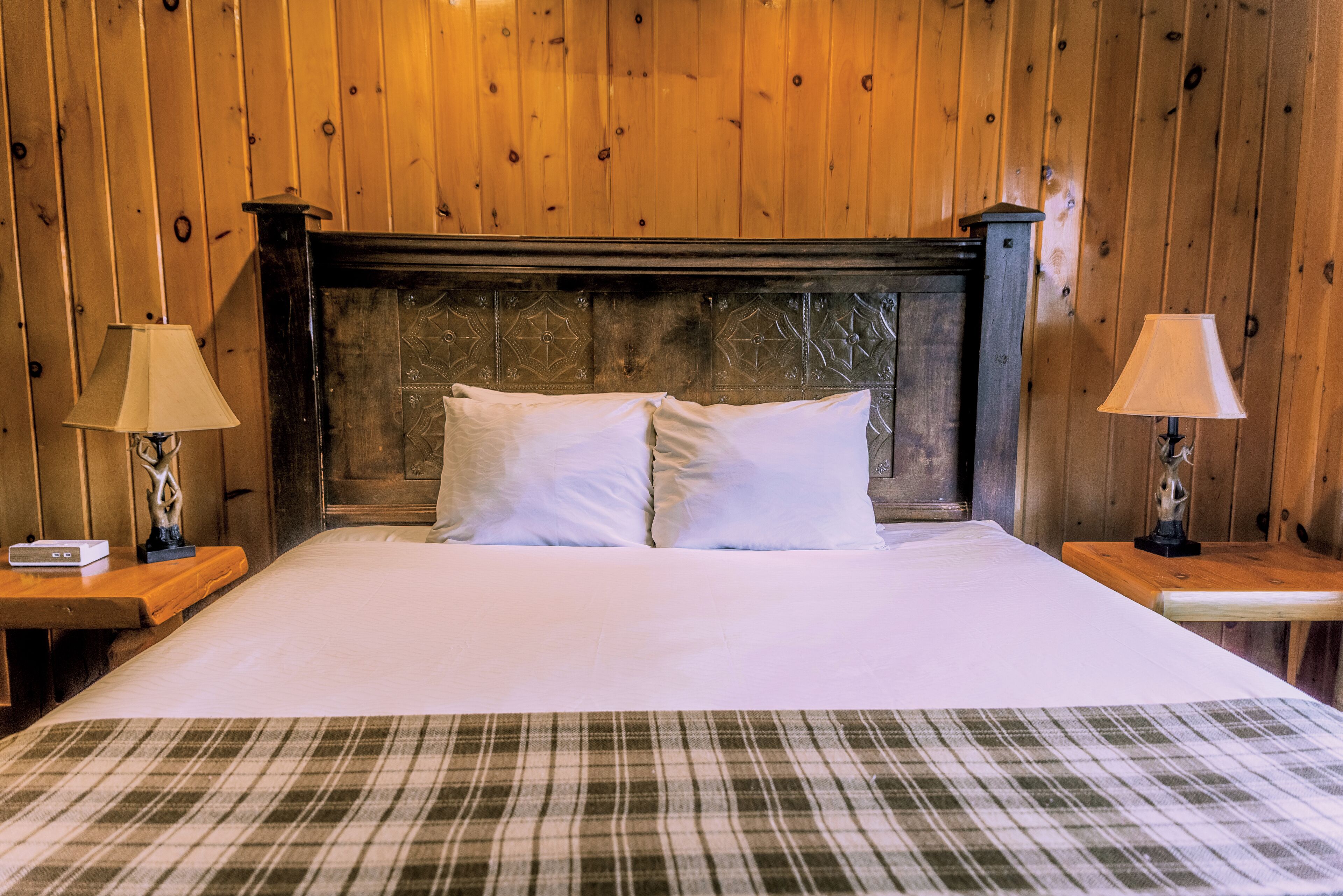 A neatly made bed with a plaid bedspread, two bedside lamps, and a wooden headboard in a room with wooden walls.