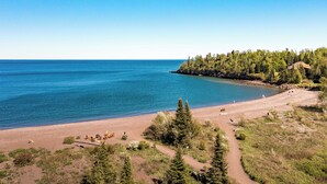 A beach with a clear view of the ocean, a sandy path, and a forested area with a small building. - Superior Shores  (Two Harbors)