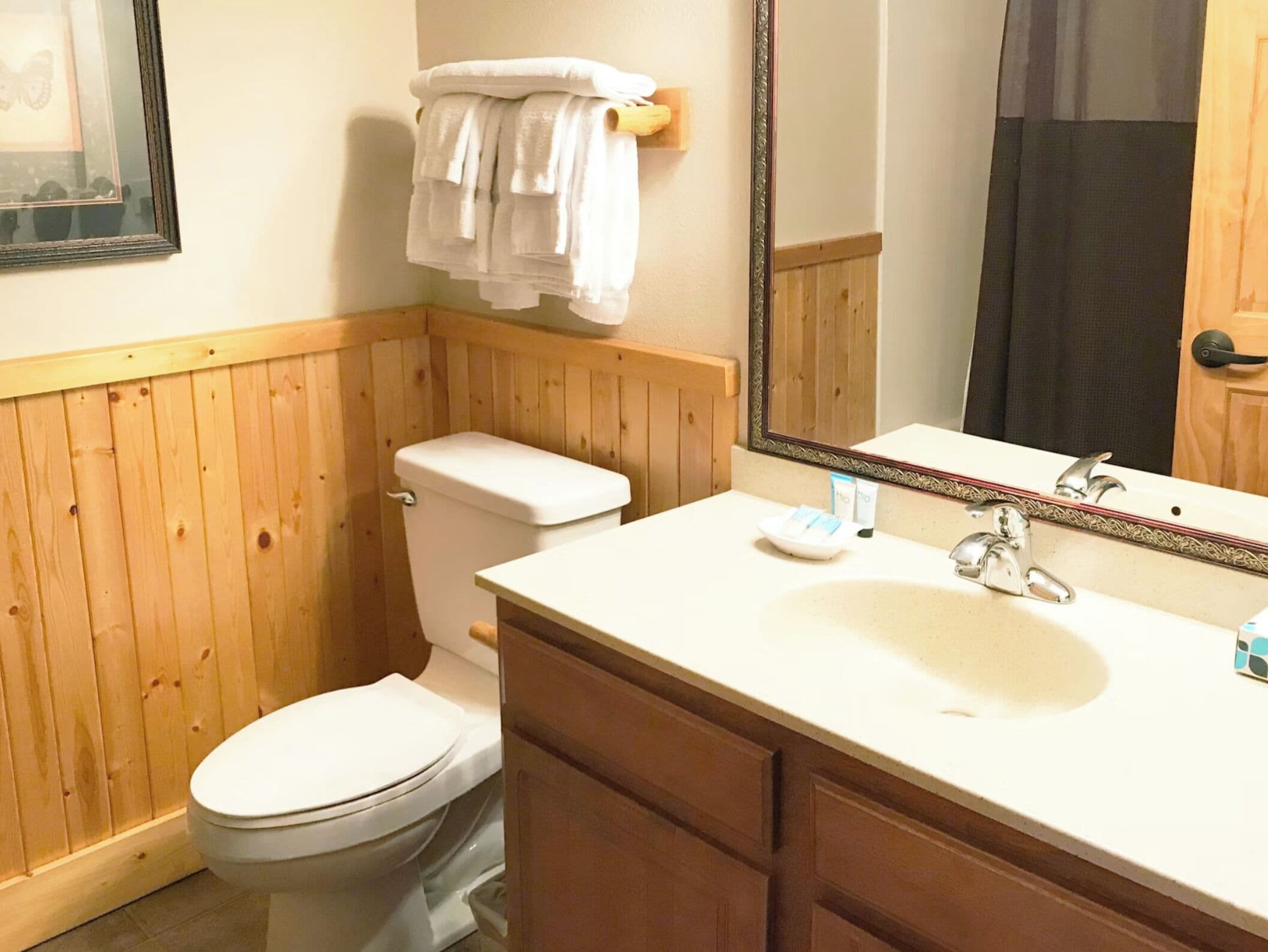 Un baño con pared de paneles de madera, un inodoro blanco, un lavamanos con espejo y un perchero con toallas dobladas.