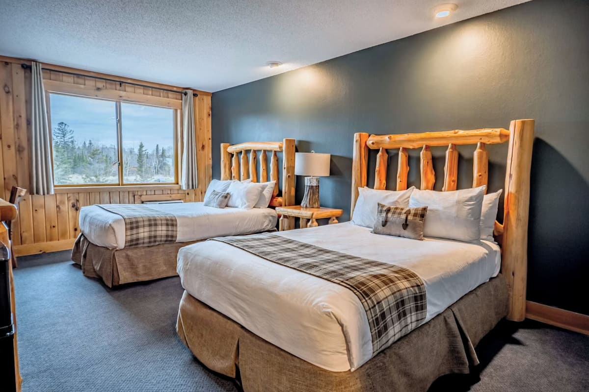 Two beds with wooden headboards, a window with curtains, and a lamp on a wooden nightstand.
