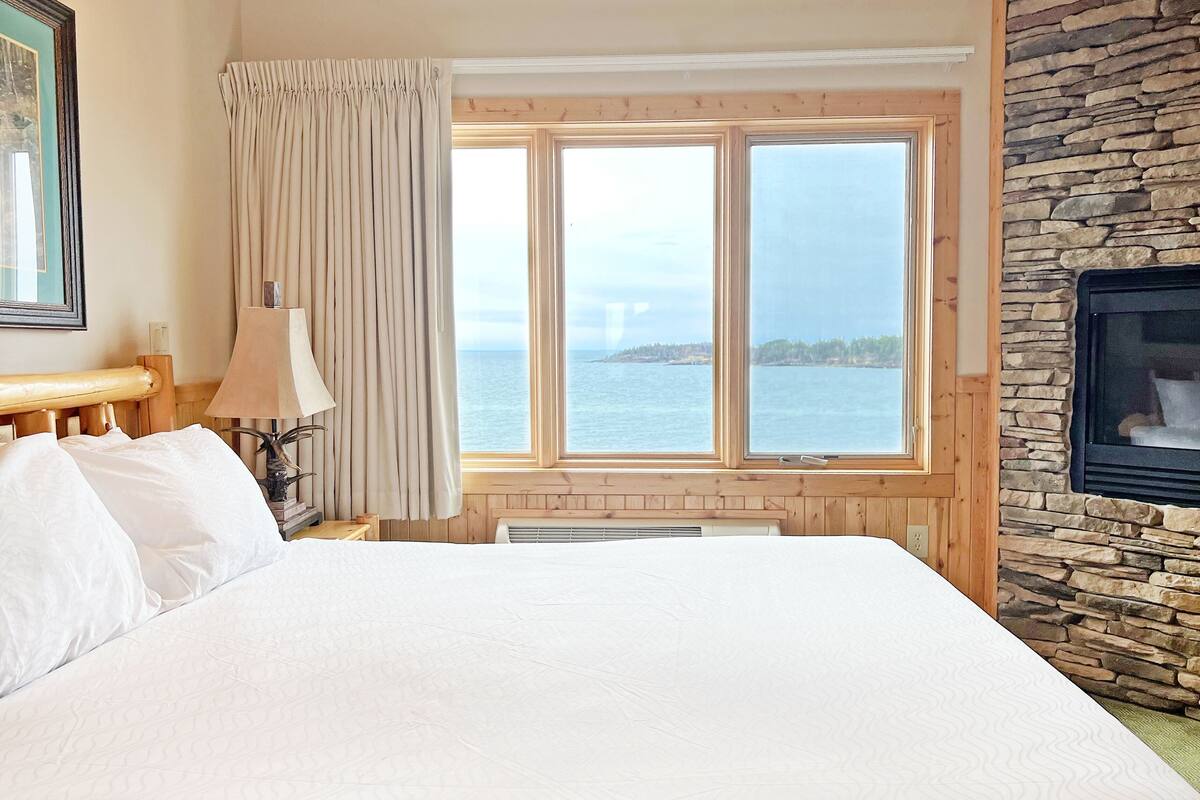 A hotel room with a large bed, a window with a view of the sea, a stone wall, and a fireplace.