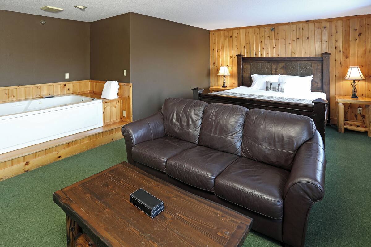A room with a wooden wall panel, a large bathtub, a leather sofa, a wooden coffee table, and a bed with two bedside tables.