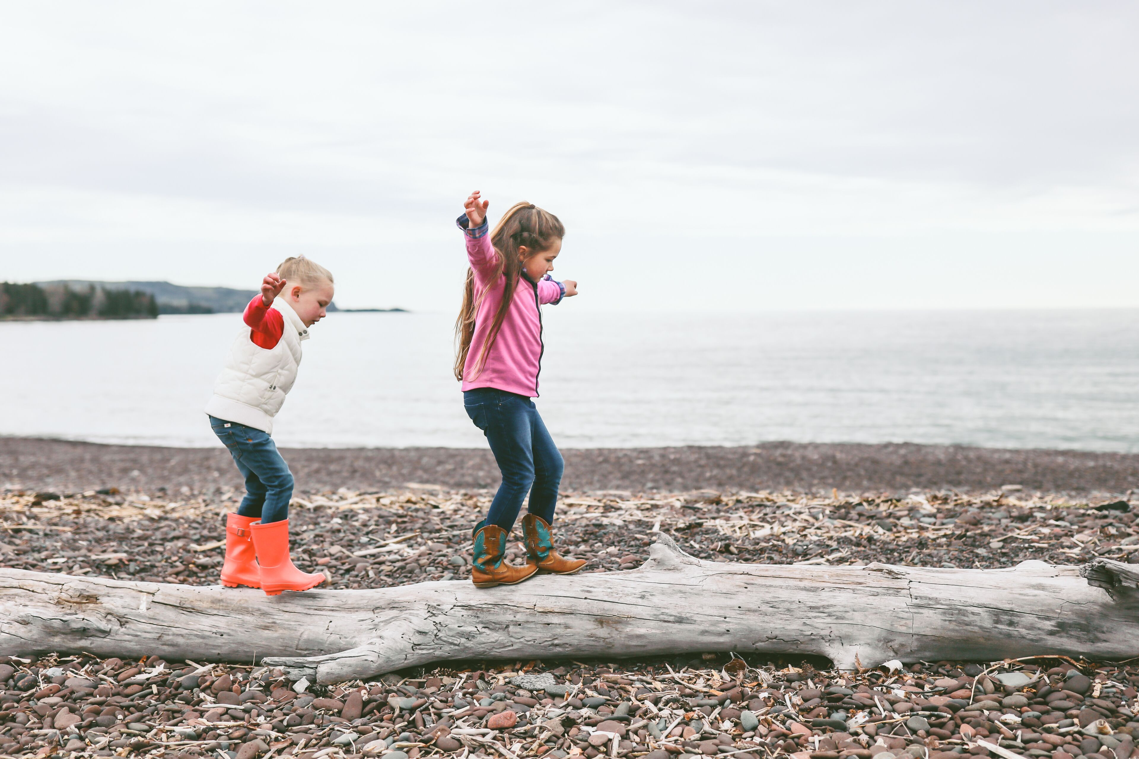 Two children playing on a log by a pebbled beach with a body of water in the background.