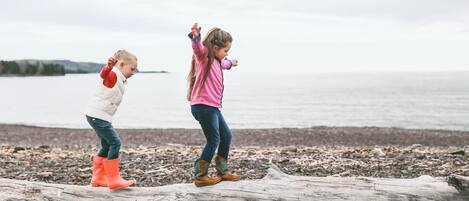 Two children playing on a log by a pebbled beach with a body of water in the background.