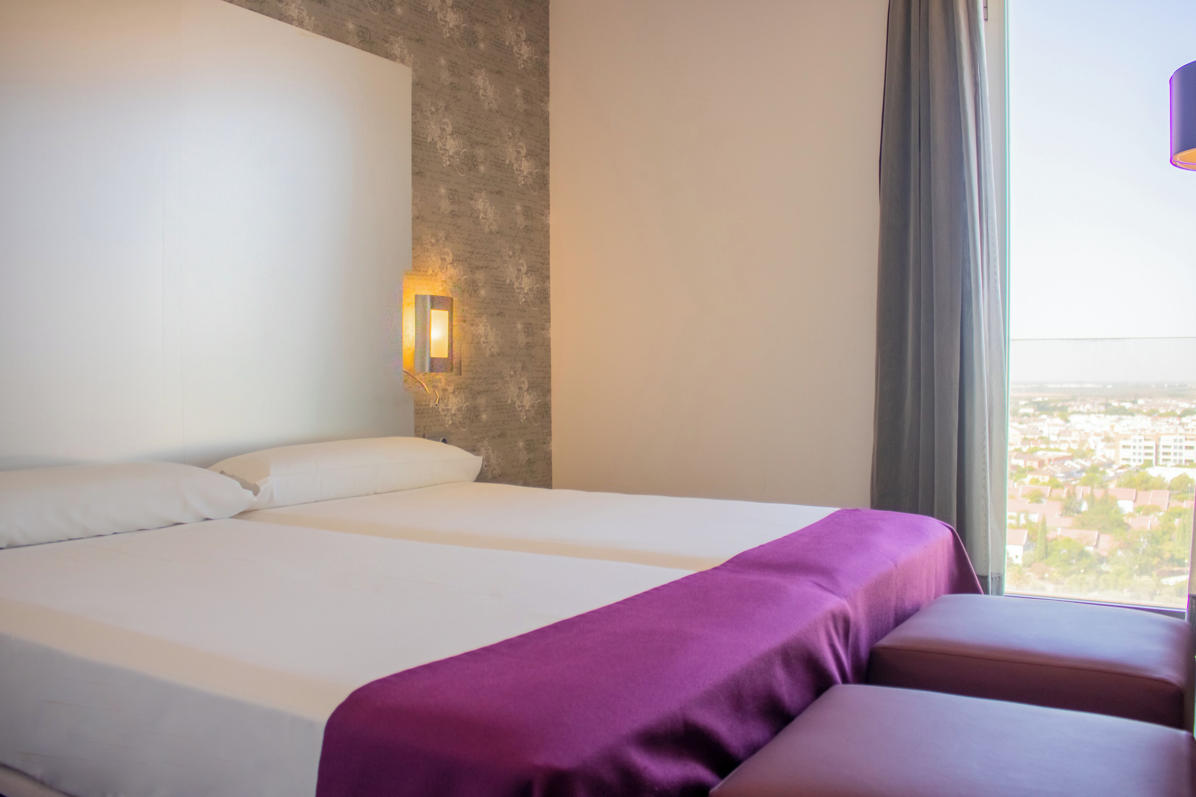A hotel room with a large bed, a small sofa, a wall-mounted light, and a window with a city view.