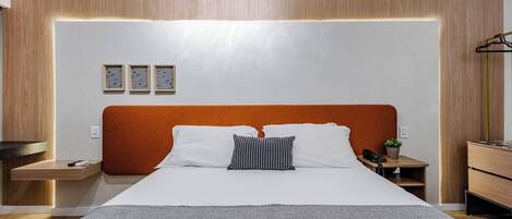 Una habitación de hotel moderna con una cama grande, cabecera de madera, mesitas de noche y un pequeño escritorio con lámpara.