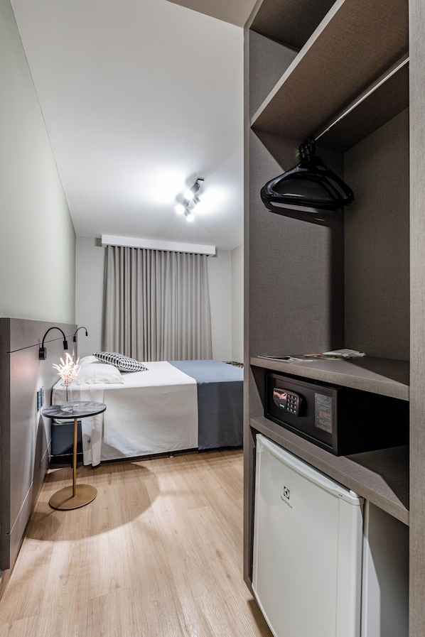 Una habitación de hotel compacta con cama, mesita de noche, lámpara y un refrigerador pequeño.