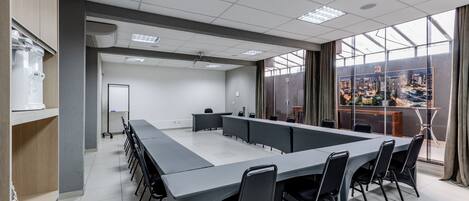 Un salón de conferencias con una larga mesa, sillas, una pizarra y vistas a un paisaje urbano a través de grandes ventanales.