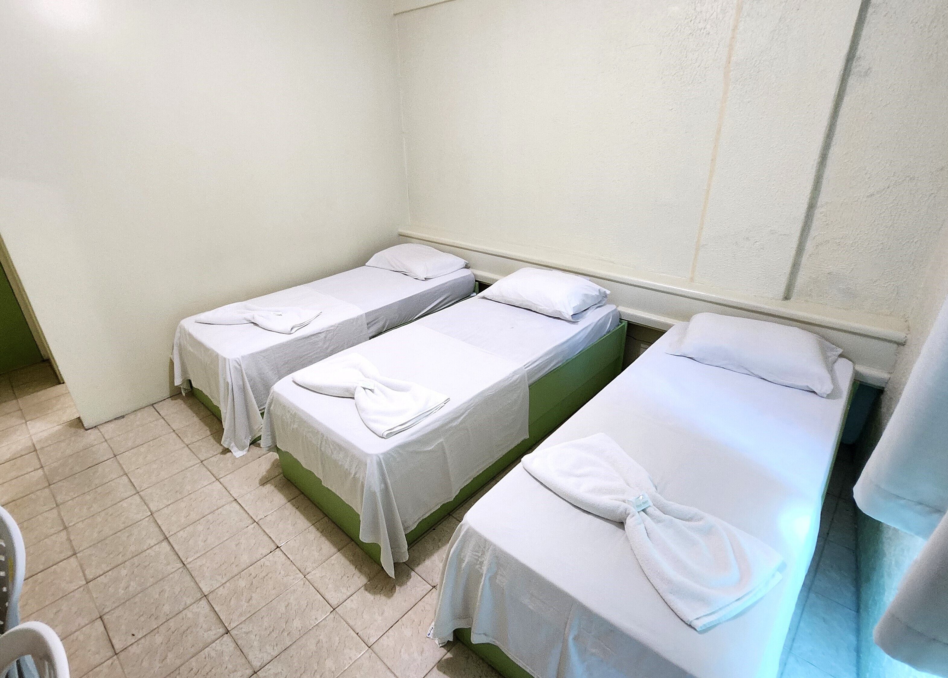 A room with three single beds, each with a white bedspread and a folded towel. The room has a tiled floor and plain walls.