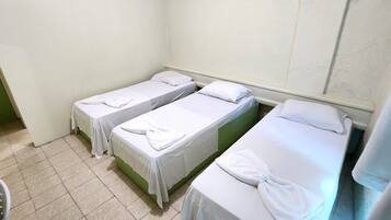 A room with three single beds, each with a white bedspread and a folded towel. The room has a tiled floor and plain walls.