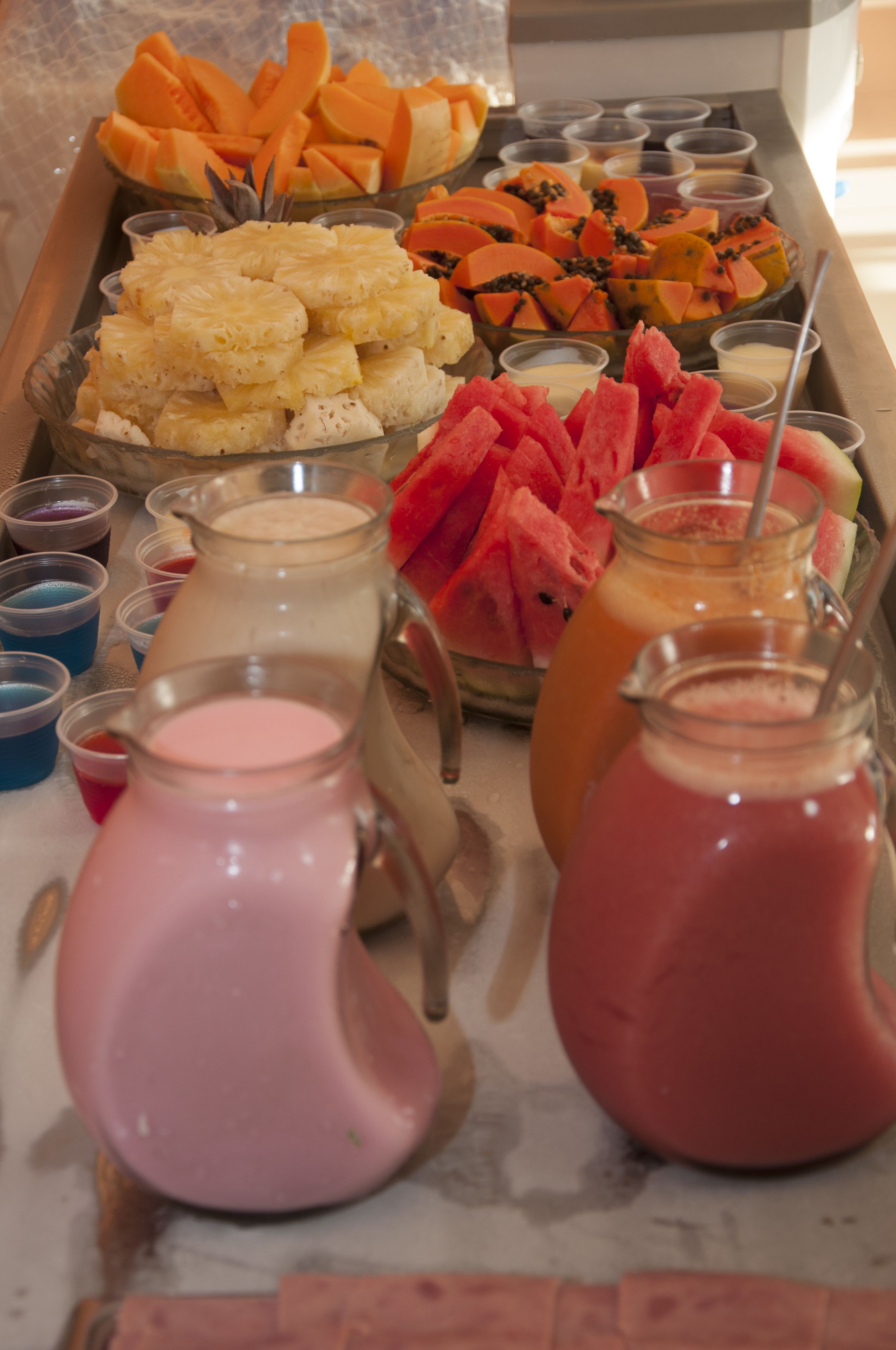 Un bufé con diversas frutas, como sandía y piña, y varias jarras de bebidas.