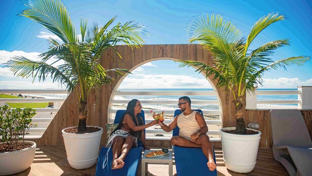 Two people sitting on lounge chairs facing a wooden archway with a view of the beach. - Opaba Praia Hotel (Ilhéus)