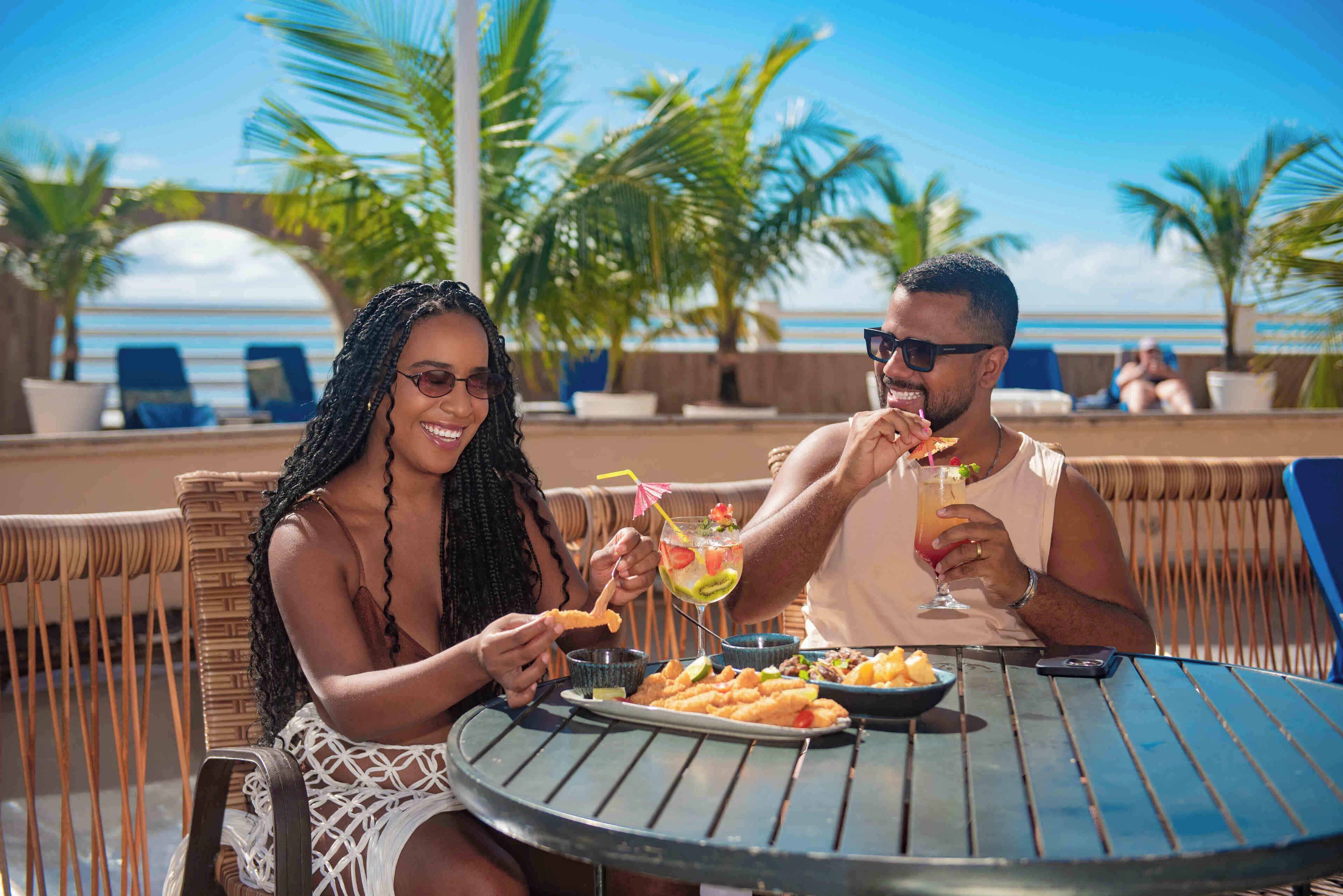 A couple dining at an outdoor table with a view of the ocean, surrounded by palm trees.
