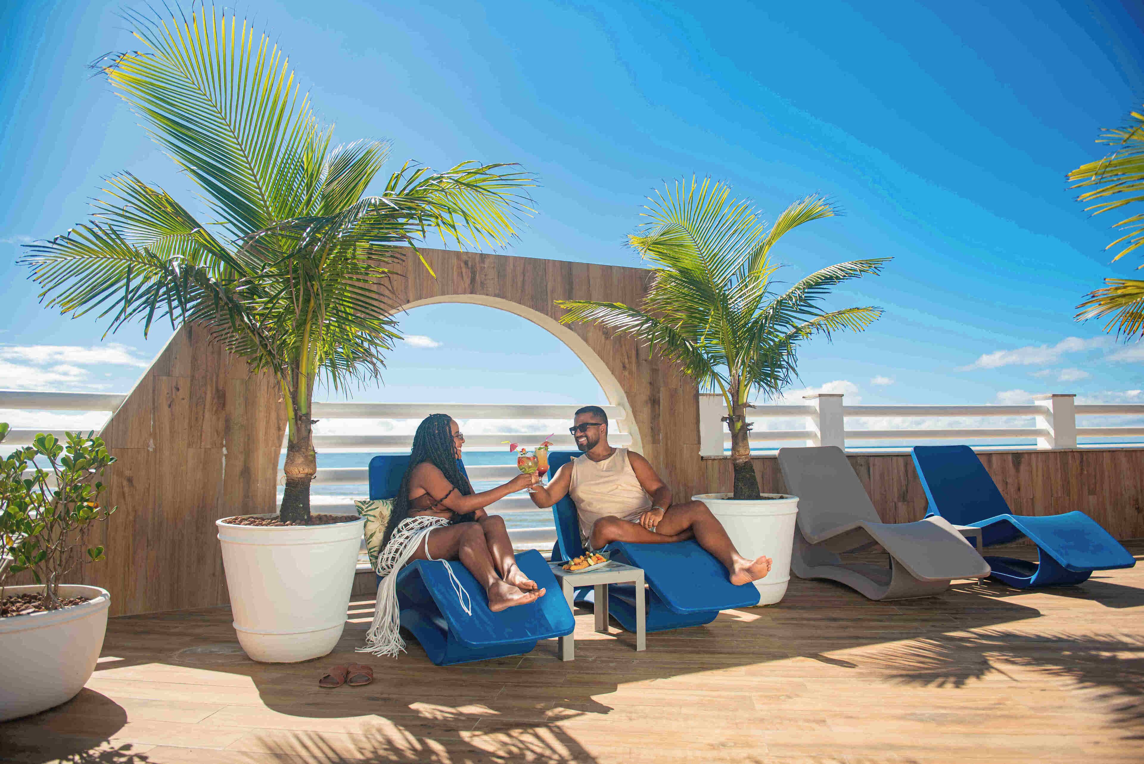 Um casal sentado em espreguiçadeiras azuis em um resort à beira-mar, com grandes vasos de plantas e um arco de madeira ao fundo.