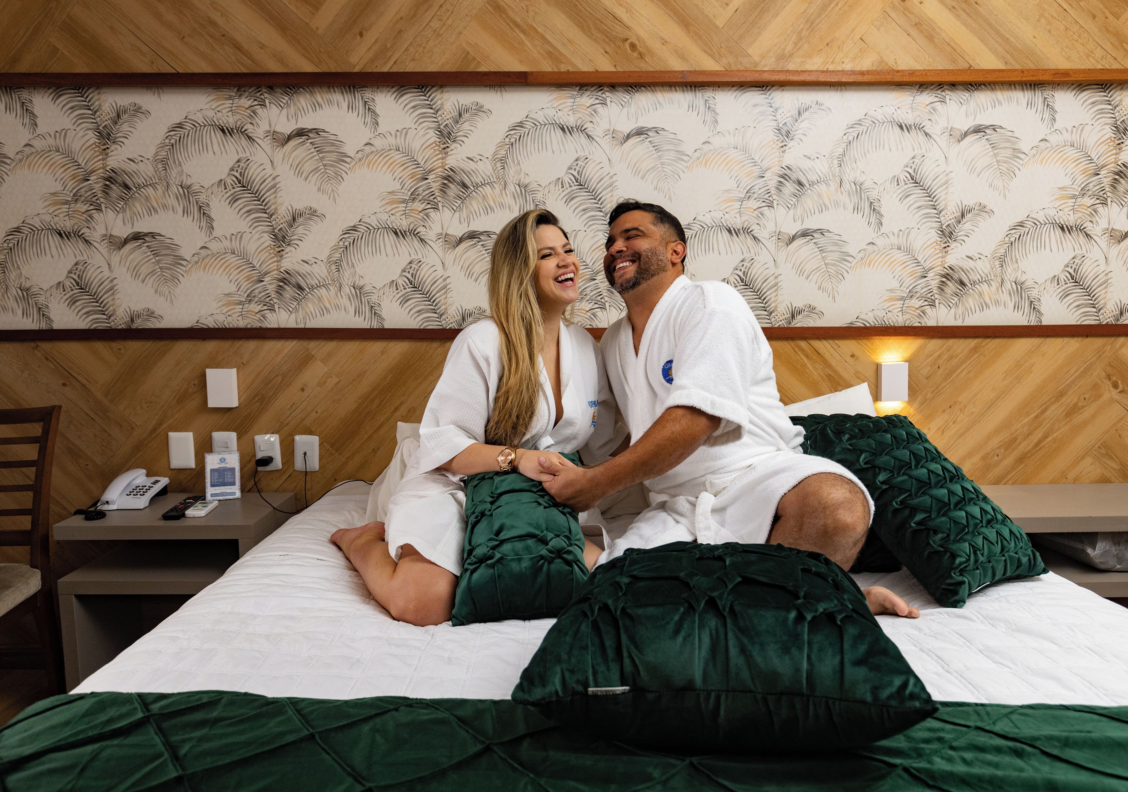 A couple sitting on a bed in a hotel room with a wooden headboard and a patterned wallpaper.