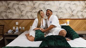 A couple sitting on a bed in a hotel room with a wooden headboard and a patterned wallpaper.