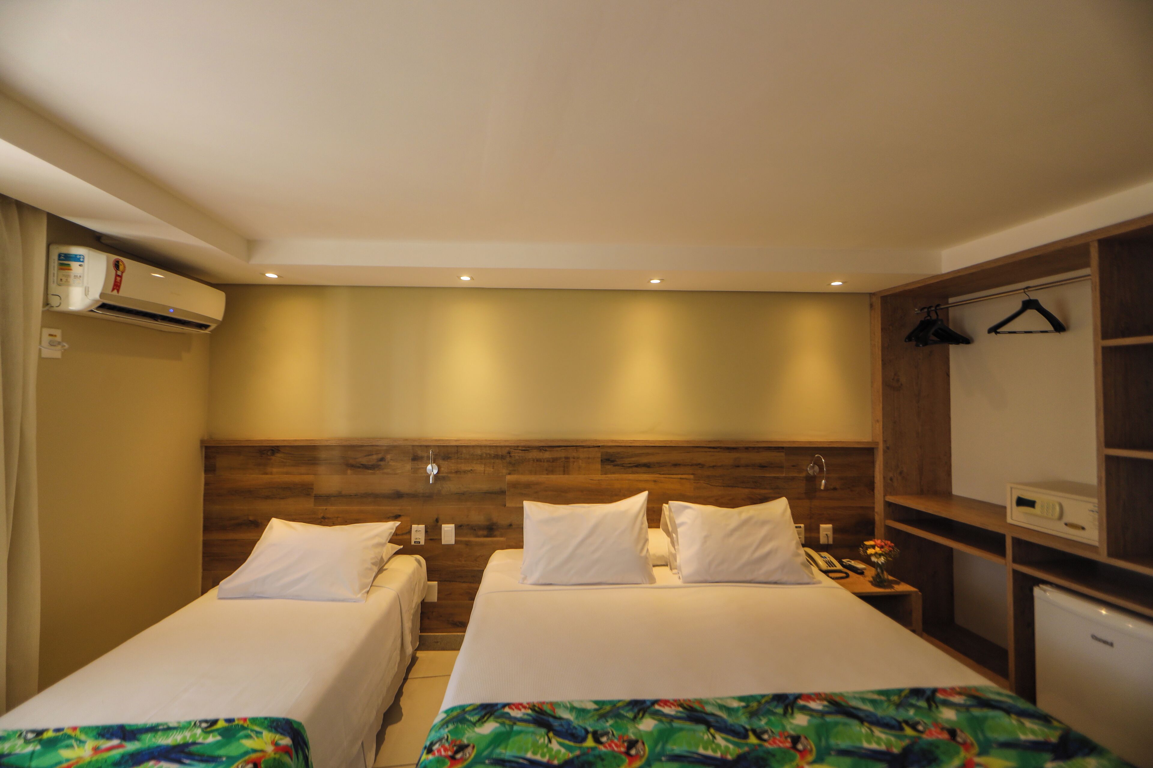 A hotel room with two single beds, a wooden headboard, a small desk with a phone and a vase of flowers, and a white air conditioning unit.