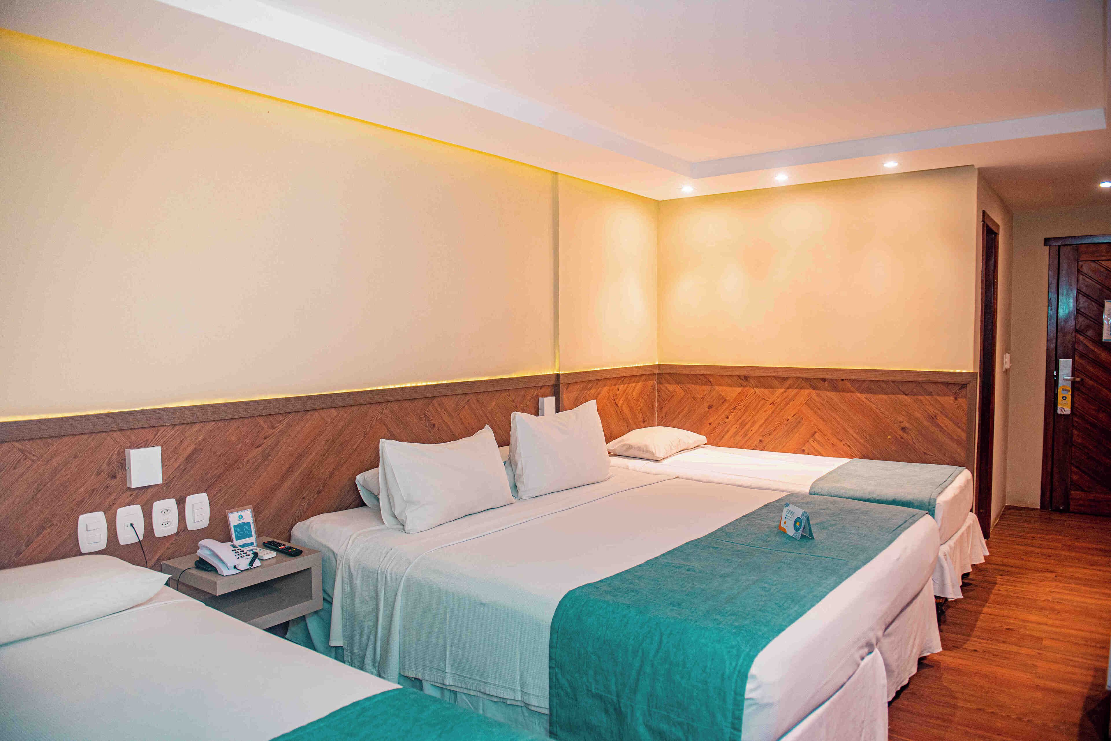 Habitación de hotel con tres camas, cabecero de madera, mesita de noche con teléfono y cargador, y una lámpara de pared.