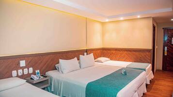 Habitación de hotel con tres camas, cabecero de madera, mesita de noche con teléfono y cargador, y una lámpara de pared.