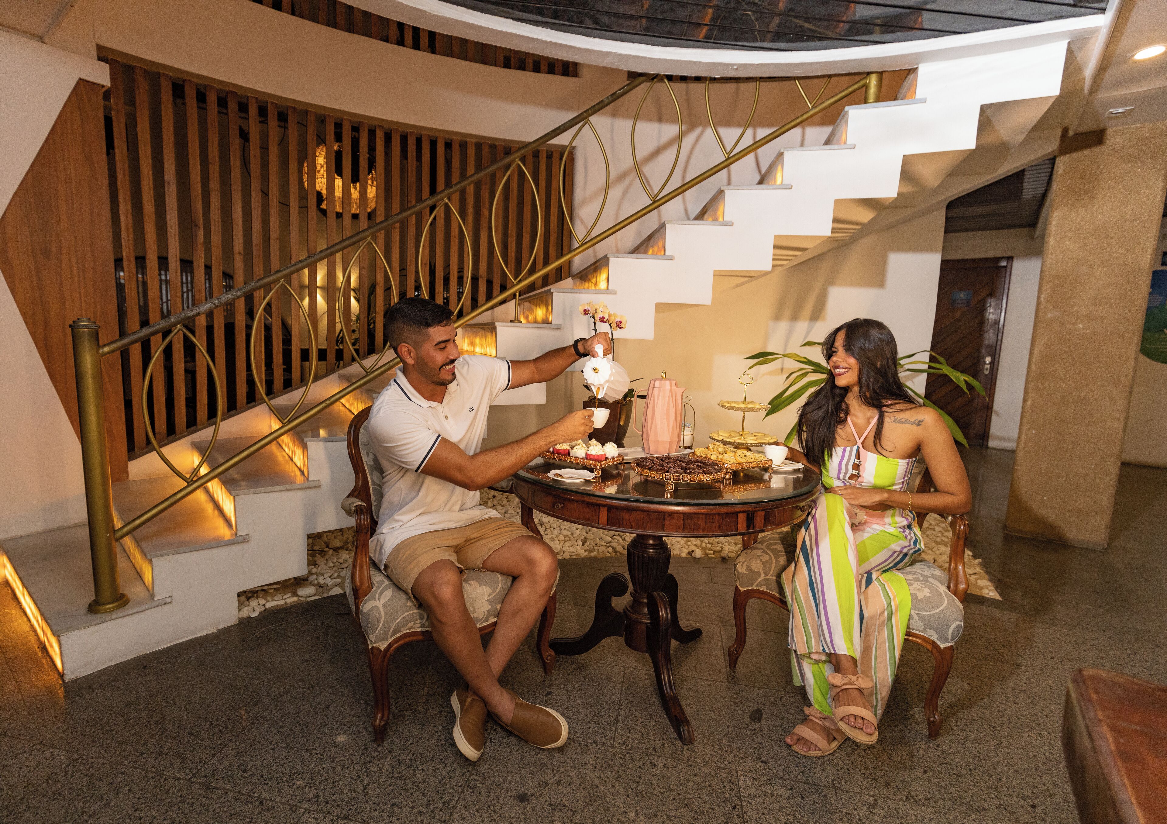 A man and a woman sitting at a table with a dessert spread in a modern interior with a staircase.