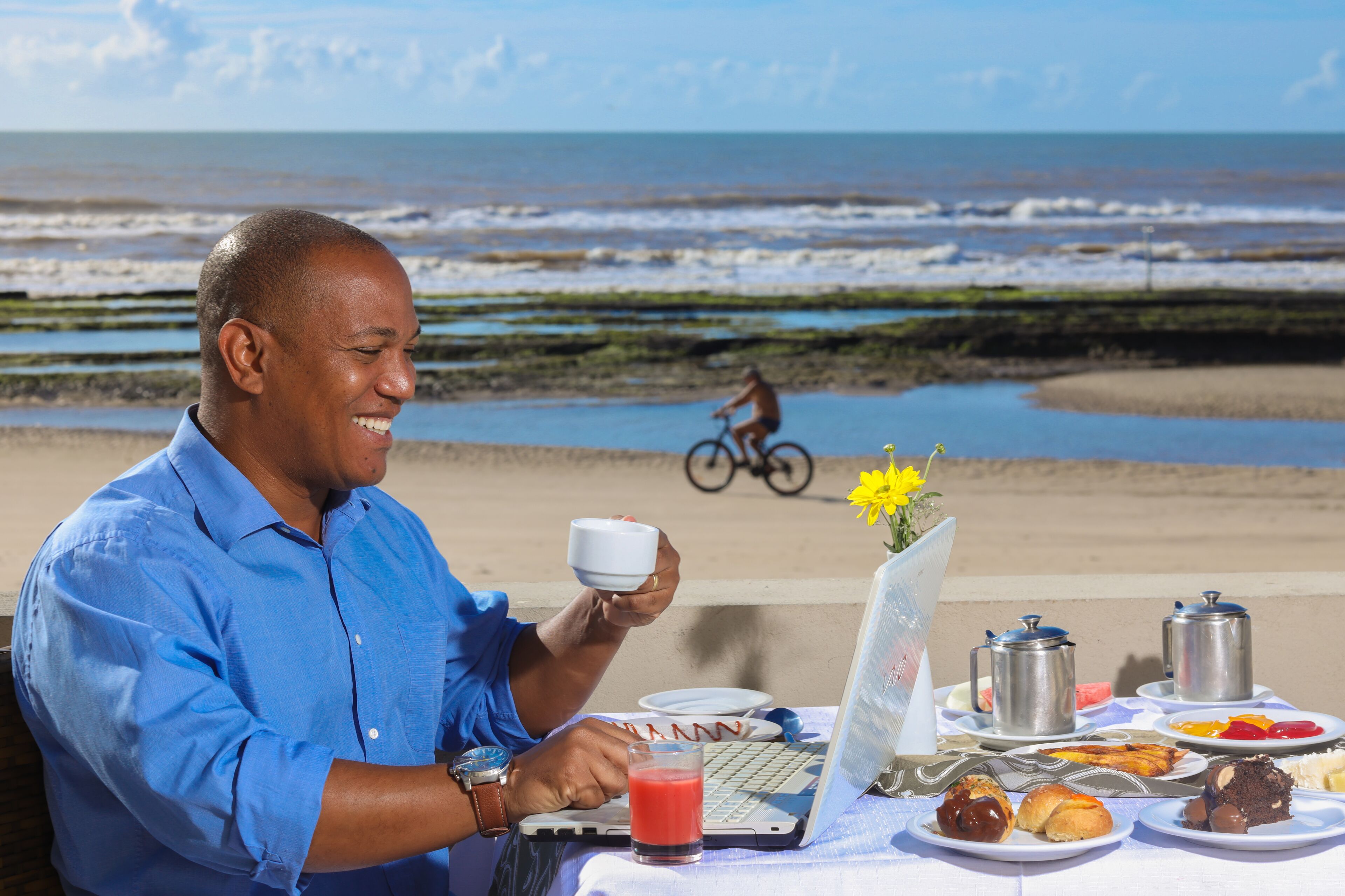 Una persona sentada a una mesa con una computadora portátil, café y comida, con vista a una playa y un ciclista al fondo.