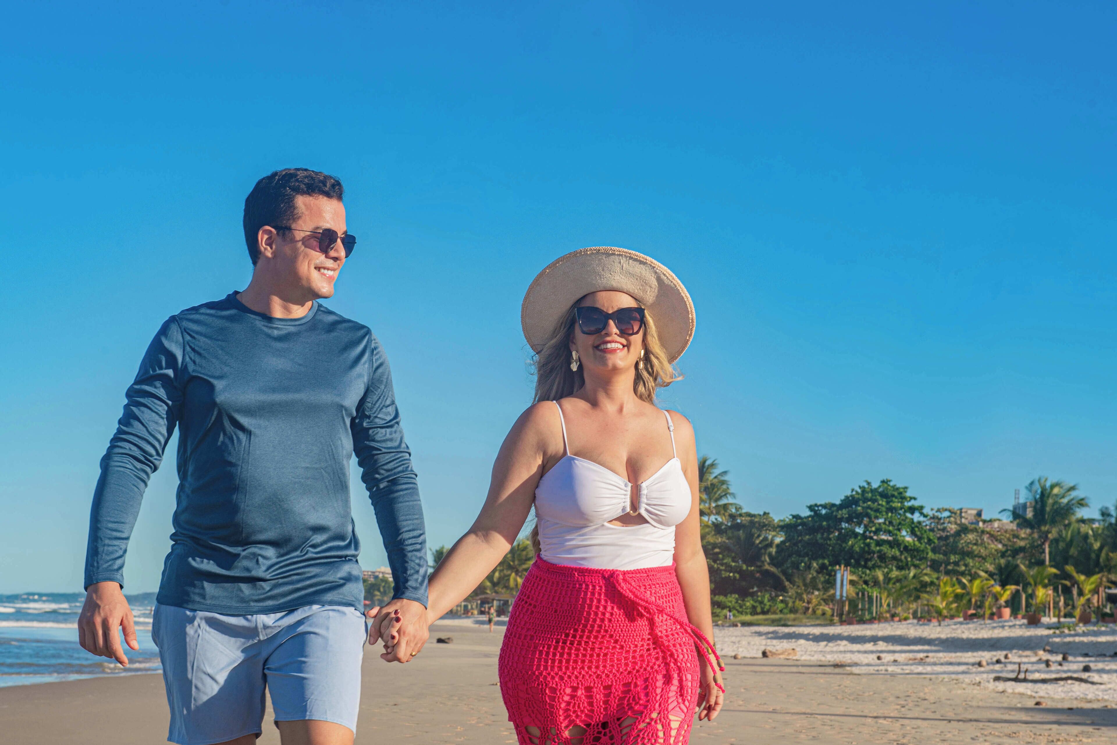 A couple walking on a beach with a clear sky and palm trees in the background.