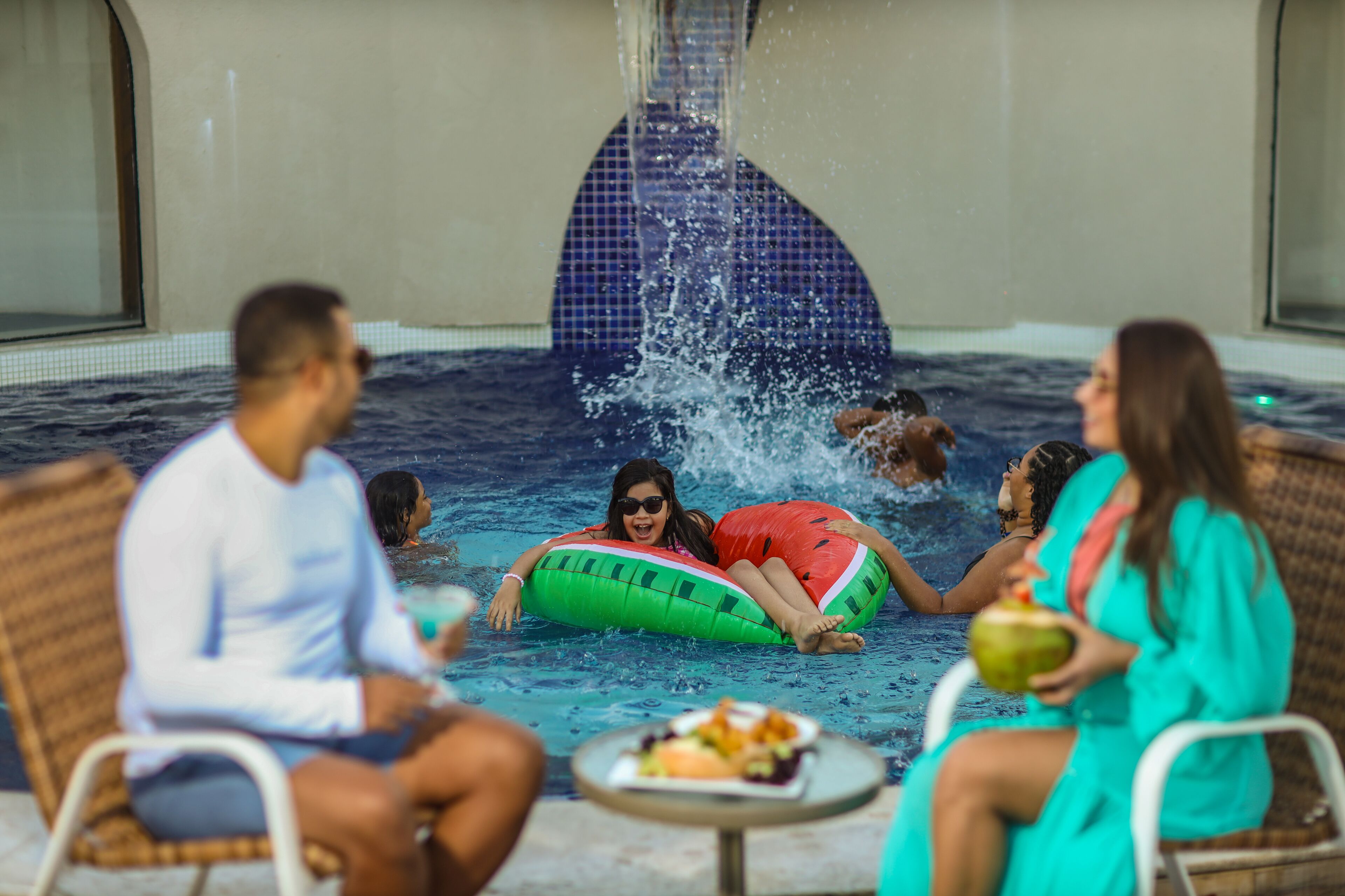 A pool area with people relaxing on chairs and floating on water toys, with a table of food in the foreground.