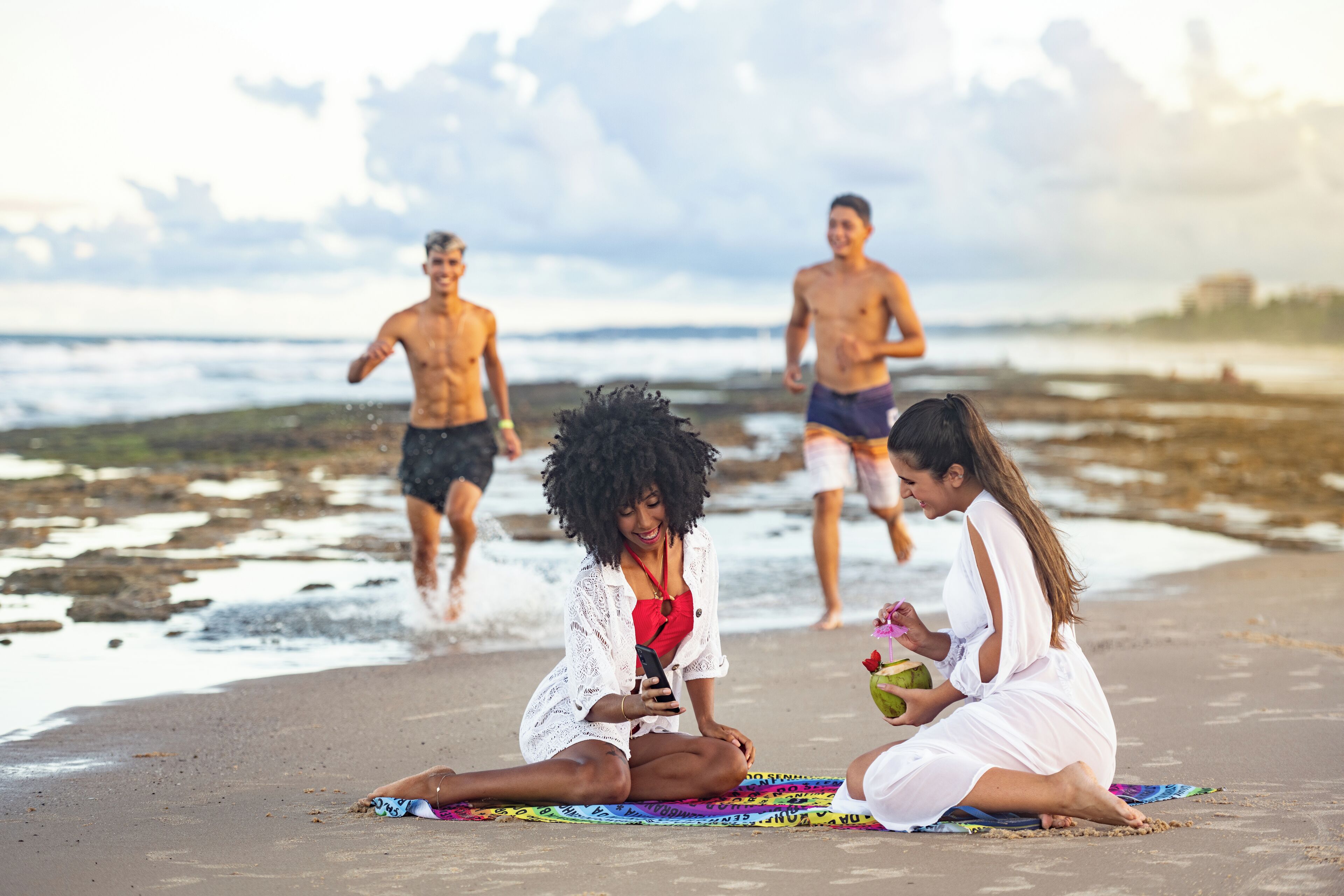A beach scene with people sitting on a colorful mat, one holding a phone and another holding a coconut.