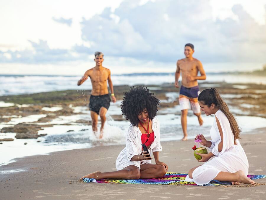 A beach scene with people sitting on a colorful mat, one holding a phone and another holding a coconut.