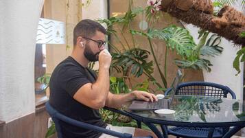 Una persona sentada a una mesa de vidrio con una computadora portátil, rodeada de follaje y una hamaca.