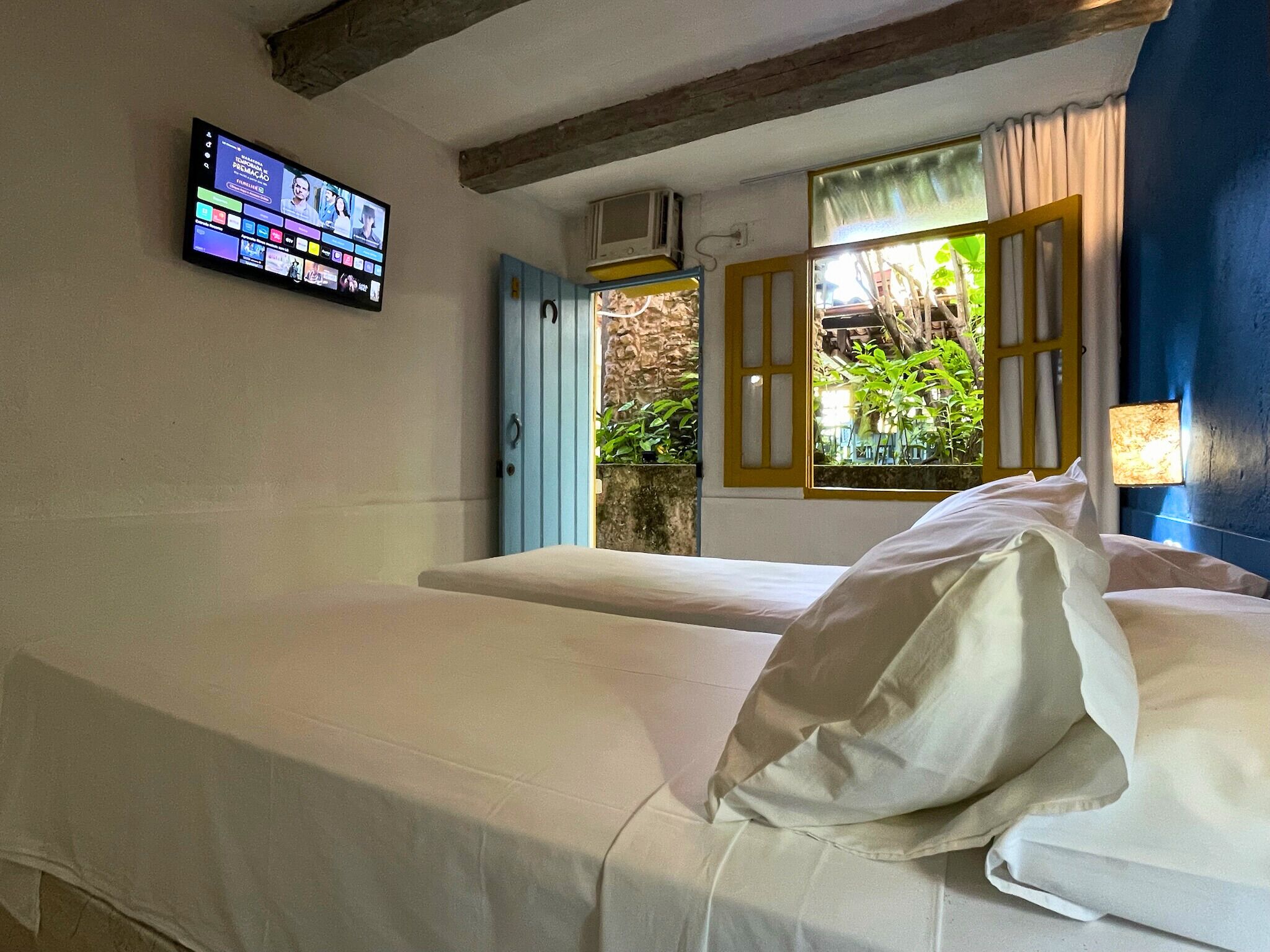 Ein Zimmer mit einem Bett, einem an der Wand montierten Fernseher und einem Fenster mit geöffneten Fensterläden.