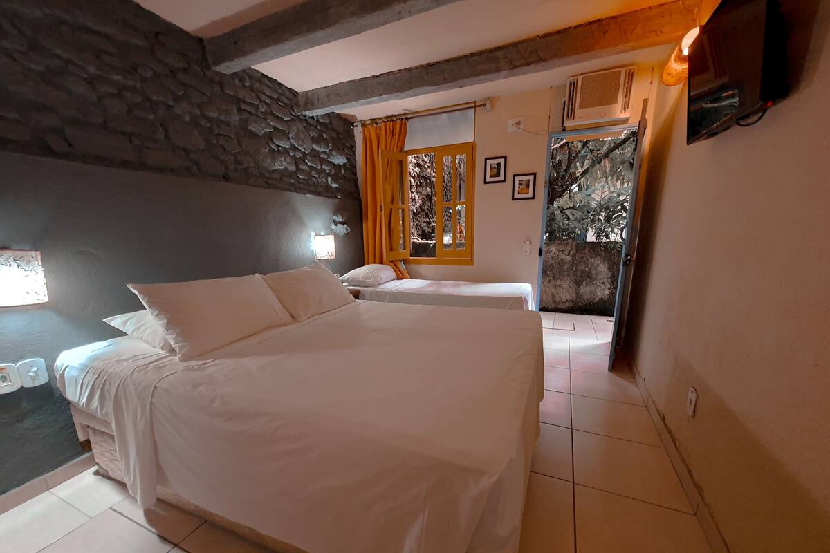 A bedroom with a large bed, a smaller bed, a stone wall, a window with curtains, and a view of trees.