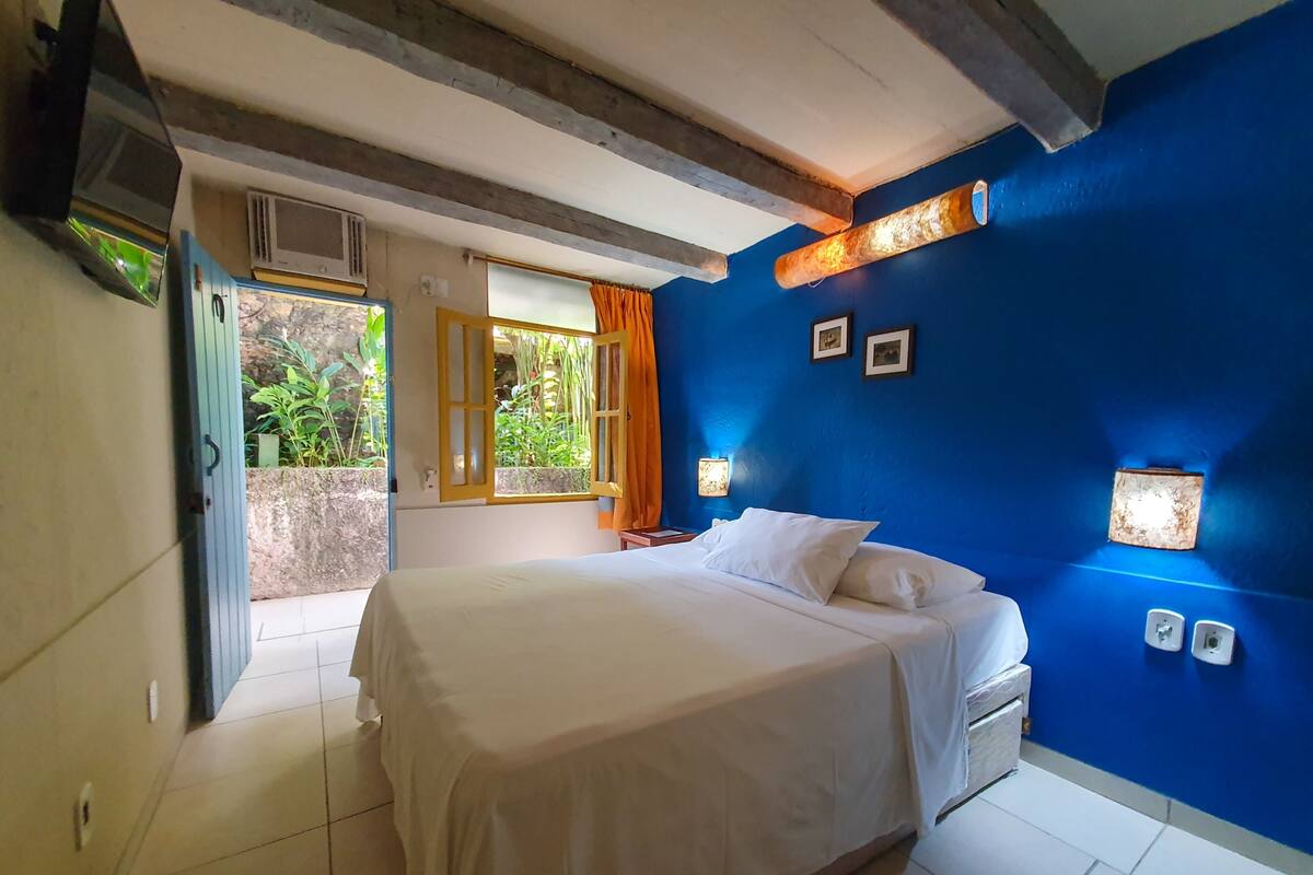 A bedroom with a bed, a window with curtains, a wall-mounted light, and a door leading outside.