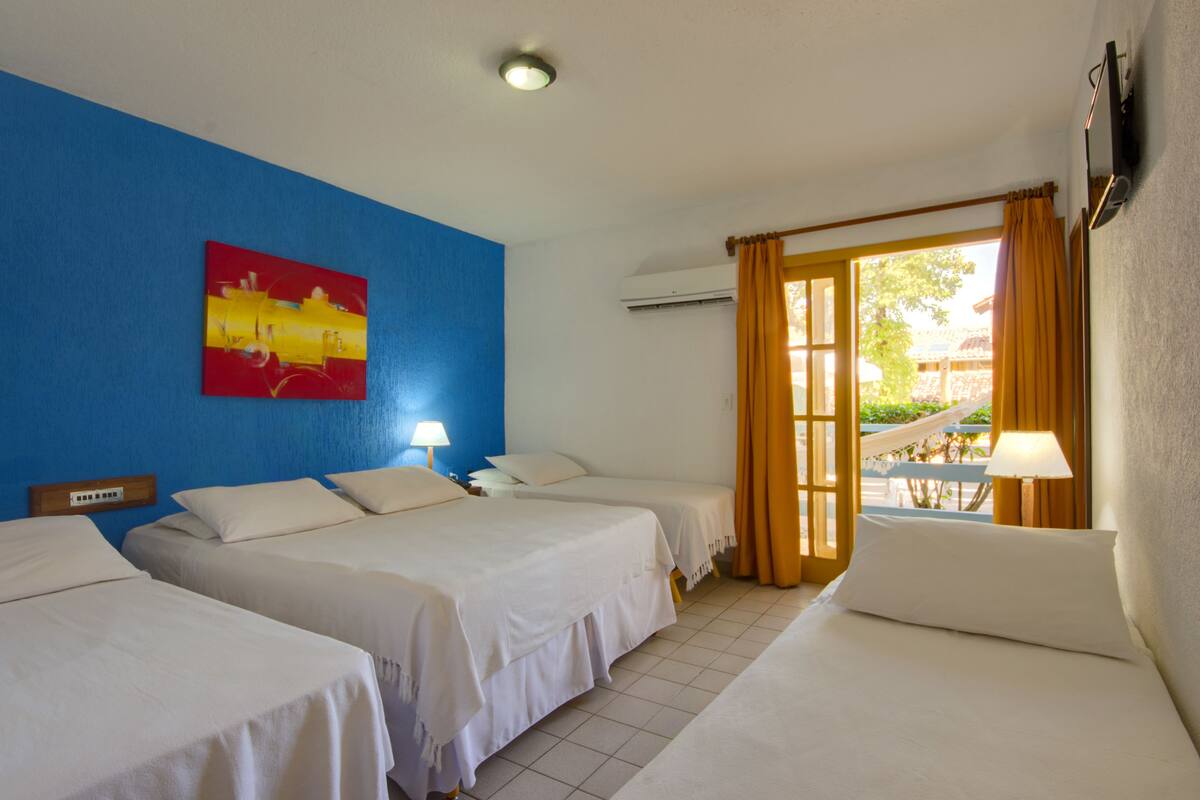 A hotel room with three beds, a blue accent wall, a painting, and a view of a garden through an open door.