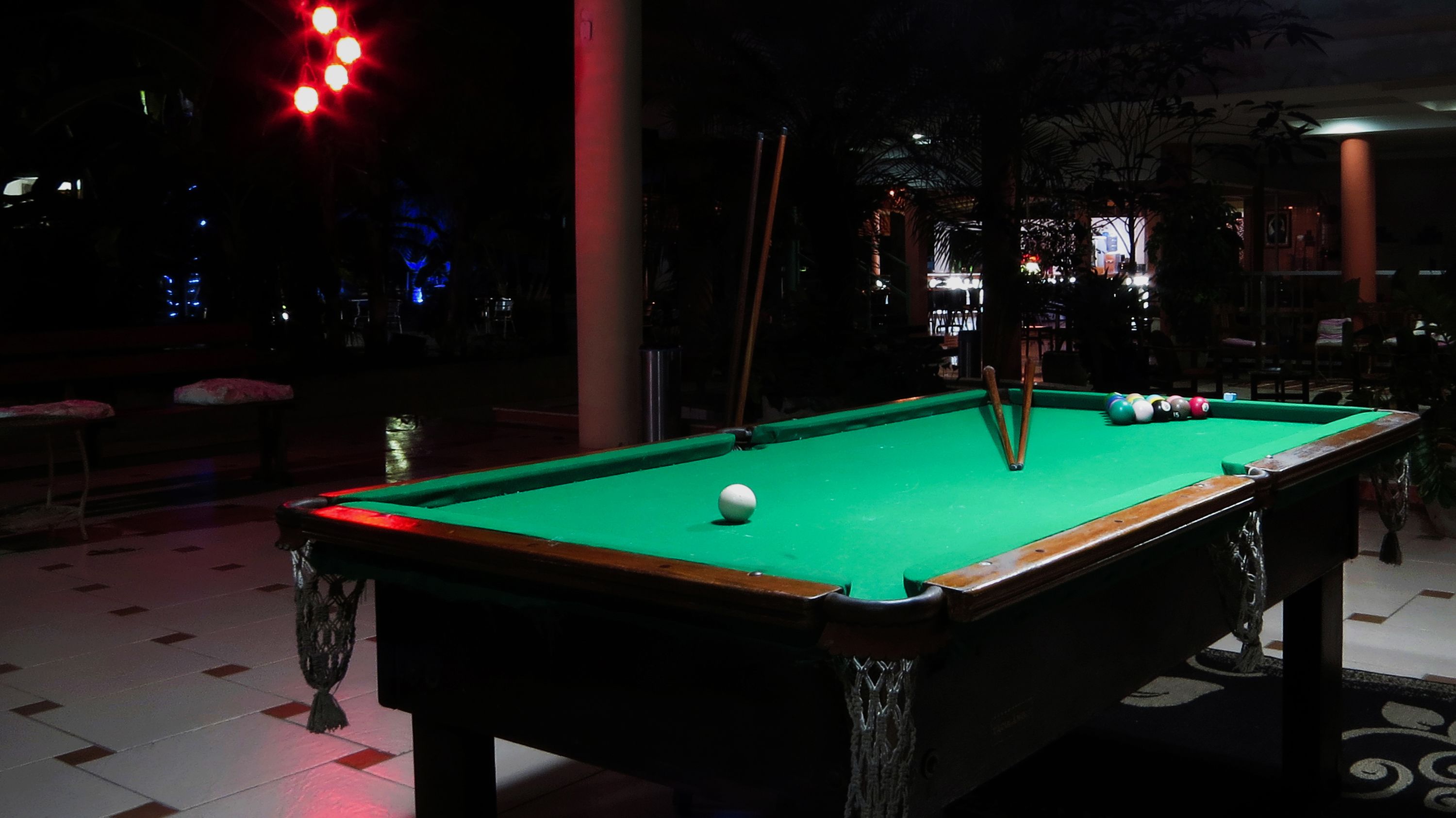 Une table de billard se trouve dans une pièce à la lumière tamisée, recouverte d’un drap de feutre vert. Elle est entourée d’un sol à motifs et de quelques chaises.