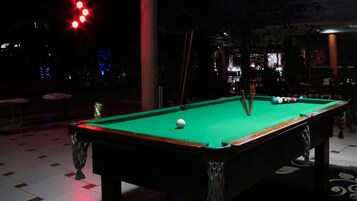 A pool table in a dimly lit room with a green felt surface, surrounded by a patterned floor and a few chairs.