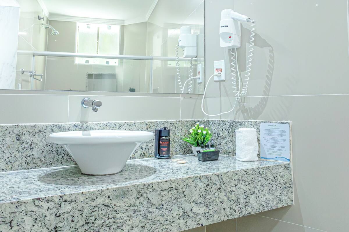 A modern bathroom with a large mirror, a sink with a marble countertop, a handheld showerhead, and a hairdryer mounted on the wall.