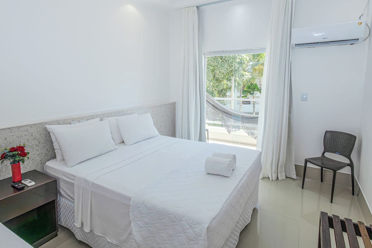 A hotel room with a bed, a chair, a nightstand, and a window with a view of greenery.