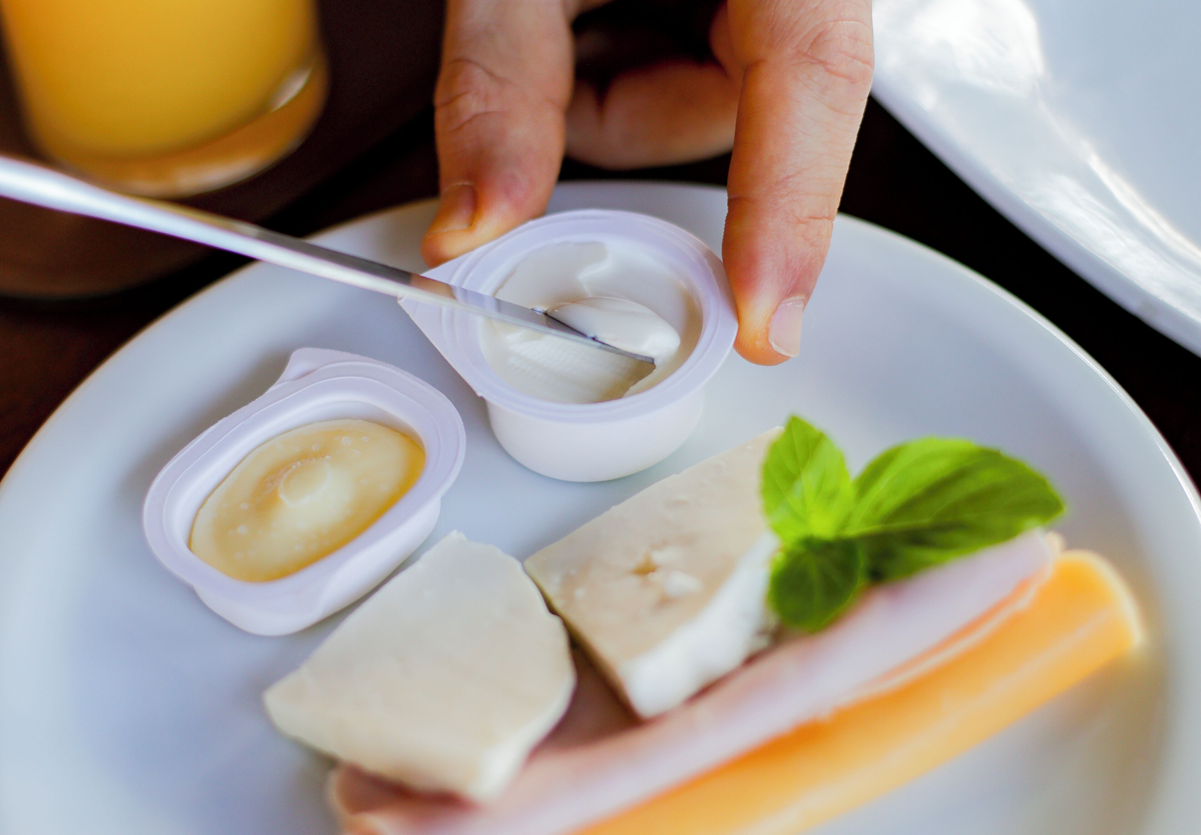 A plate with a slice of ham, cheese, and a small container of sauce, with a hand holding a spoon.