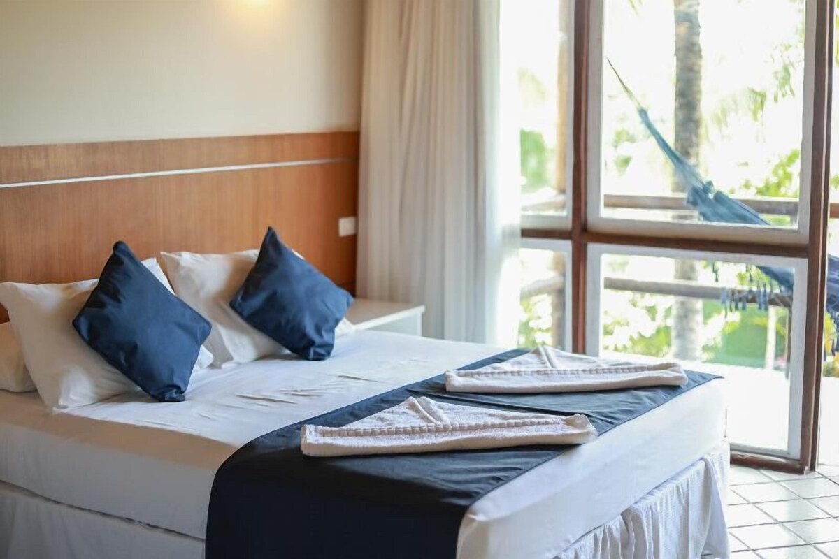 Habitación de hotel con cama, almohadas, una manta y vistas al exterior a través de amplias ventanas.