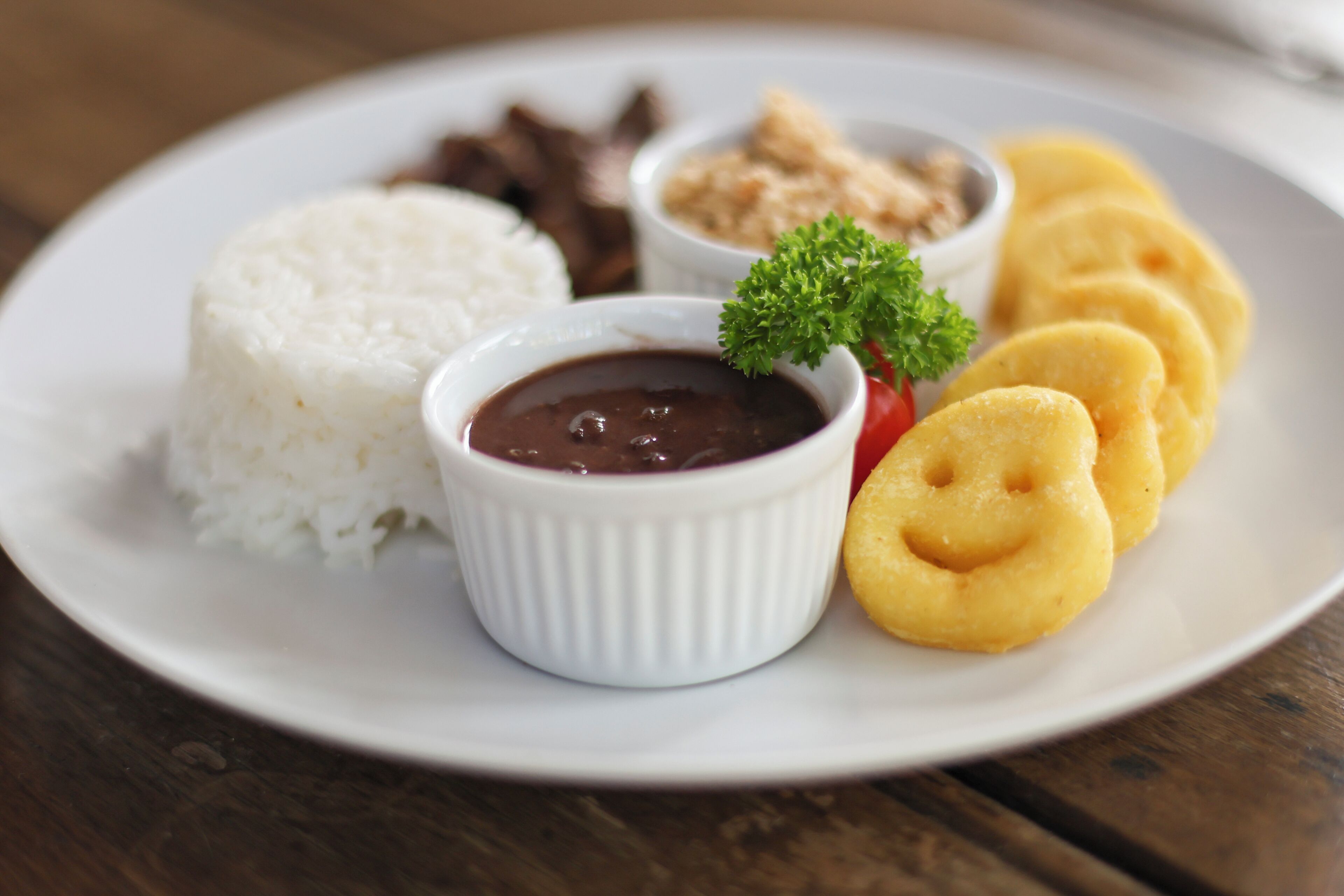 A plate with white rice, a small bowl of black sauce, and yellow fried items, possibly fritters, on a wooden table.