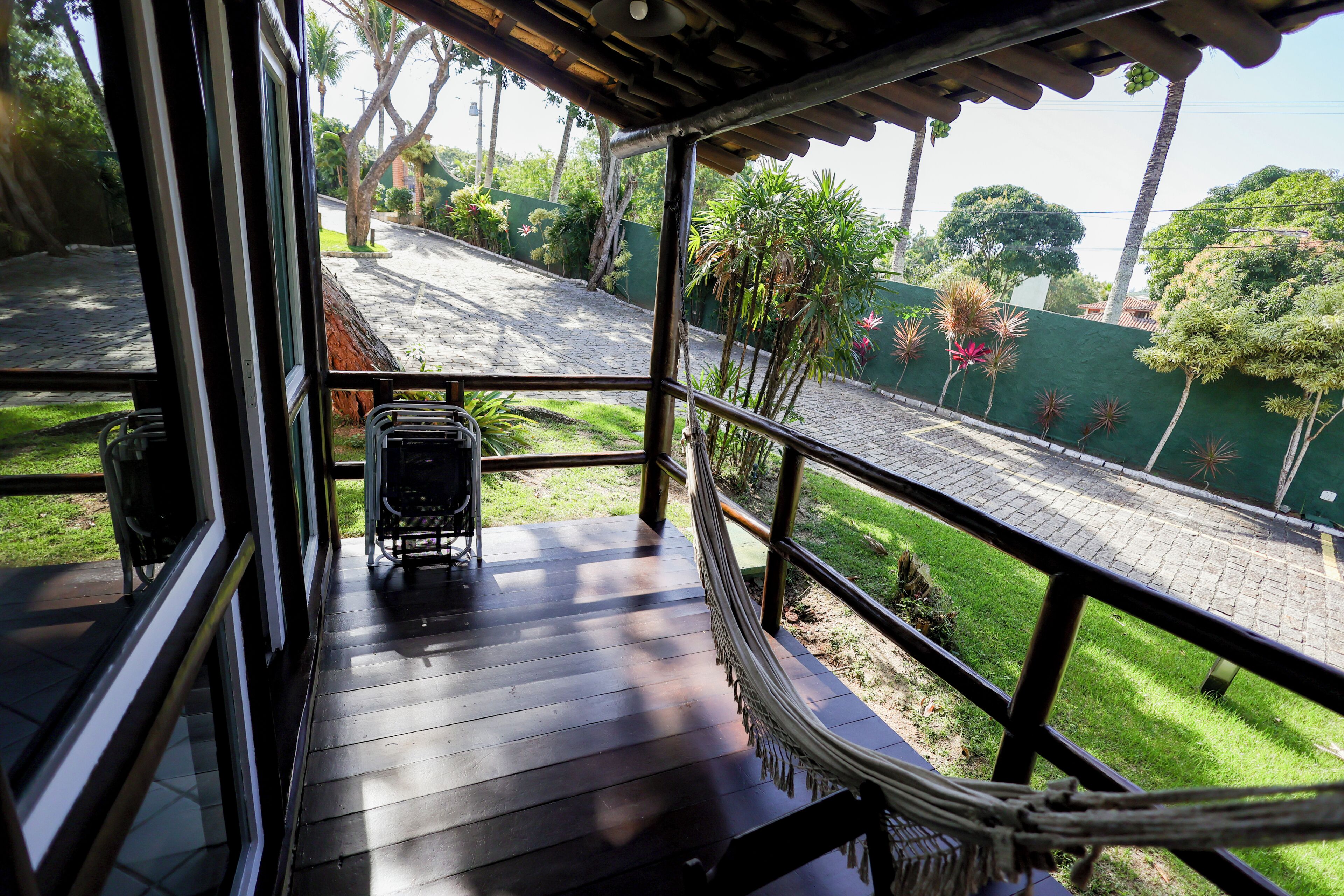 A wooden deck with a metal railing overlooking a paved area with greenery and a building in the distance.