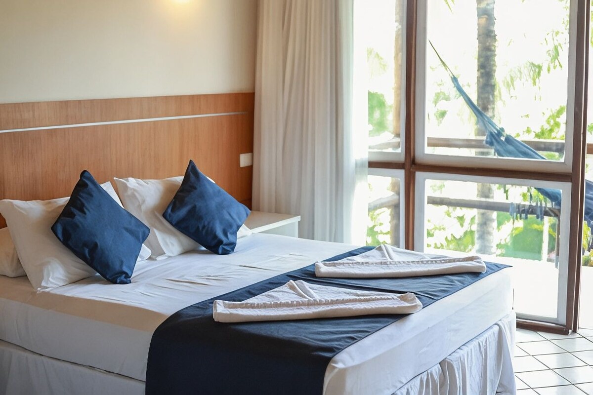 Habitación de hotel con cama, ropa de cama blanca, almohadas azules, cabecero de madera y vistas a zonas verdes a través de amplios ventanales.
