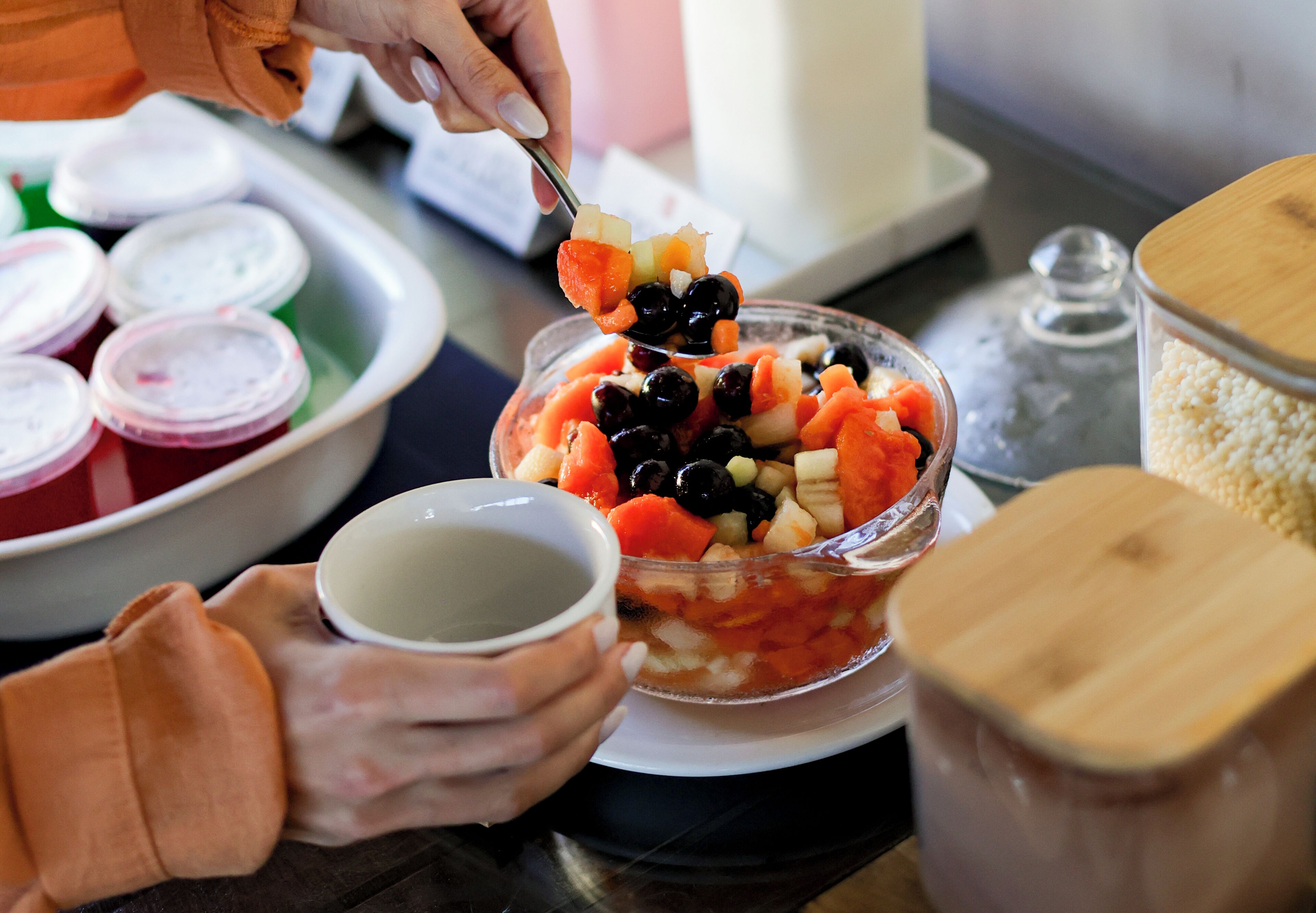 A person preparing a fruit salad with a spoon, a bowl of fruit, a cup, and a plate with food containers in the background.