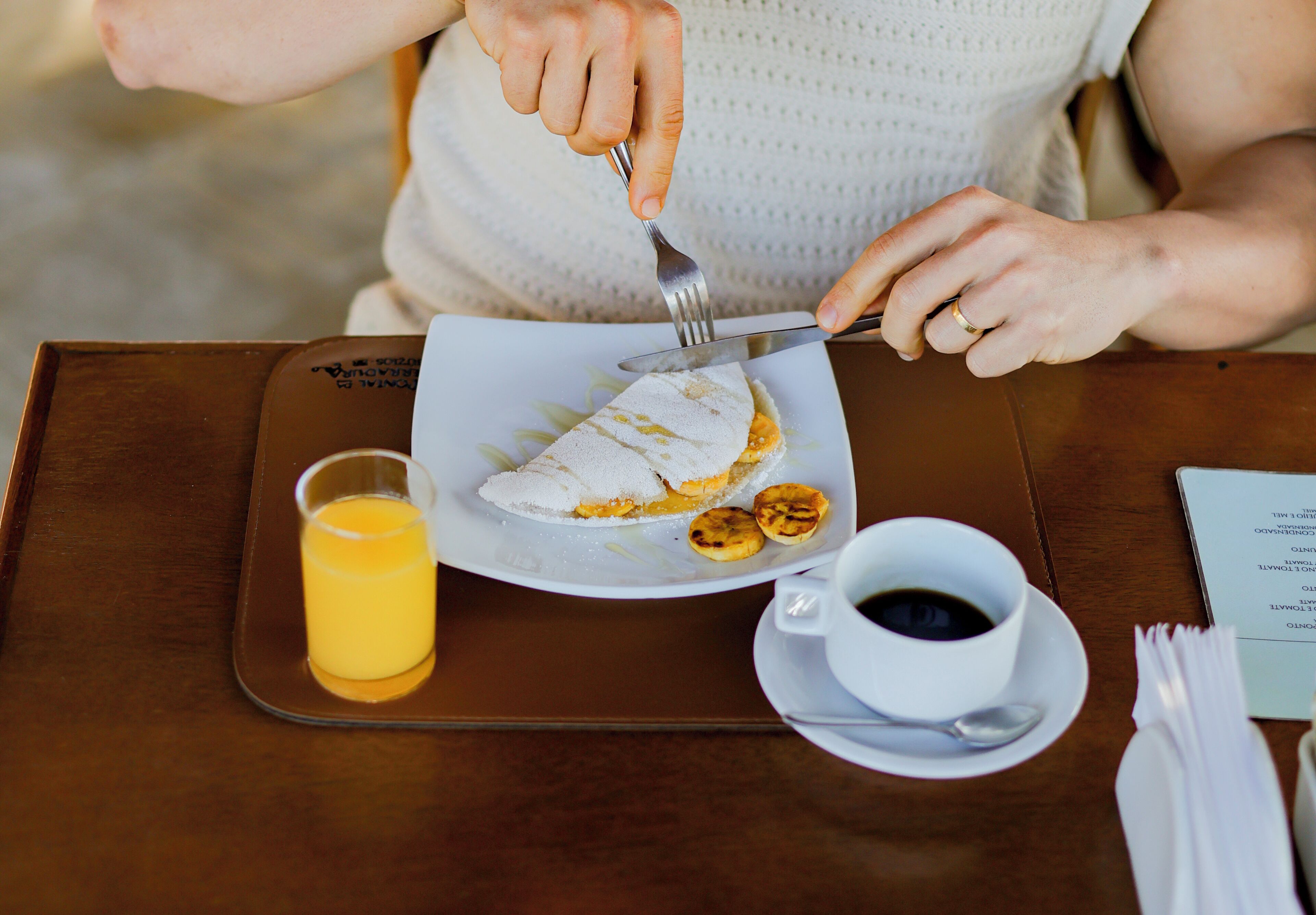 Une personne coupe un sandwich sur une assiette blanche à l’aide d’une fourchette et d’un couteau. On voit également un verre de jus d’orange et une tasse de café sur une table en bois.