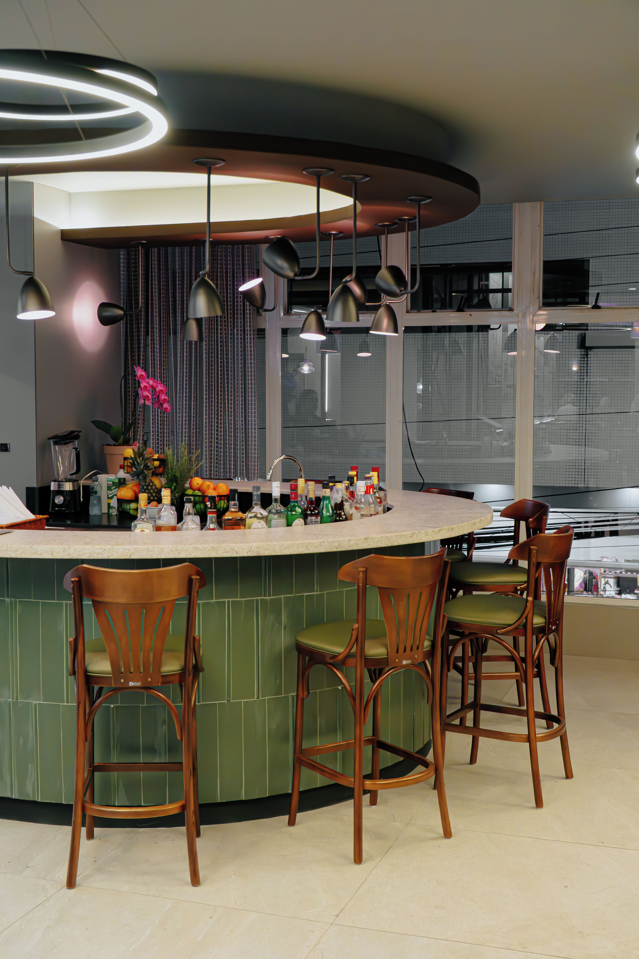 A modern kitchen with a curved island, bar stools, pendant lighting, and a view of the city through large windows.