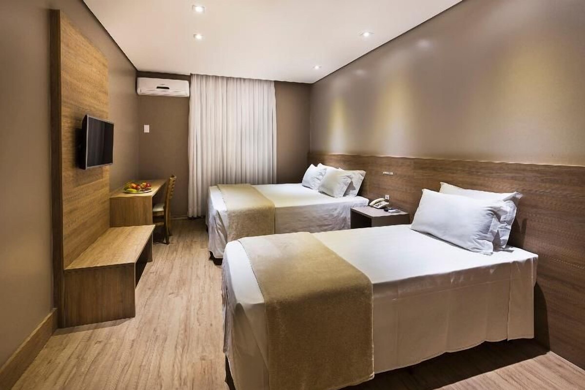 A hotel room with two beds, a desk, a chair, a TV, and a window with curtains.