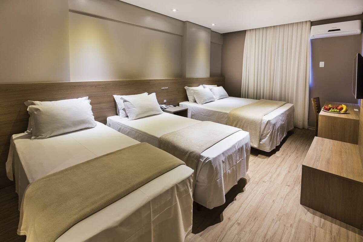 Hotelkamer met drie bedden, een houten hoofdeinde, een bureau met een schaal fruit en een wandairconditioner.