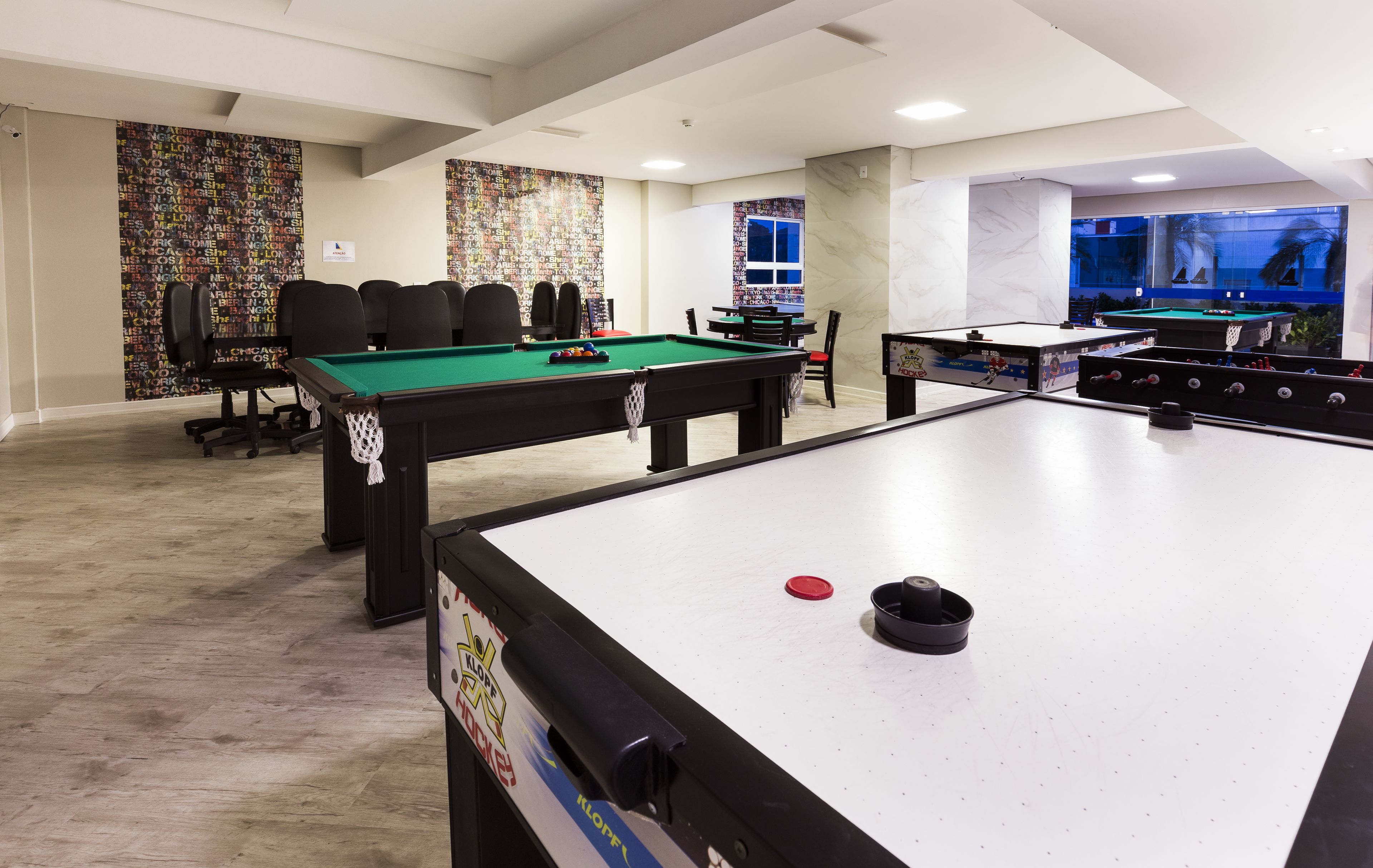 A pool table, a foosball table, and a billiards table in a room with a modern design, including a wall with city names and a window with a view.