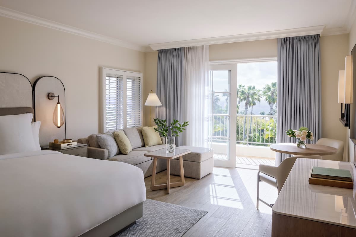 A hotel room with a large bed, a sofa, a small table with a vase of flowers, a desk with a chair, and a balcony with curtains.